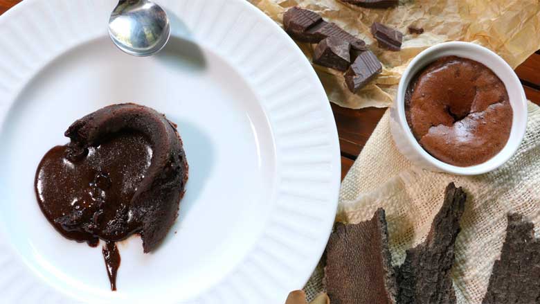 A top shot of a broken molten chocolate cake with chocolate flowing out. The cake lies on a white plate with a spoon showing. With another chocolate lava cake in a white ramekin, chocolate pieces on brown parchment paper, and wood bark in the background - Chocolate Lava Cake Recipe