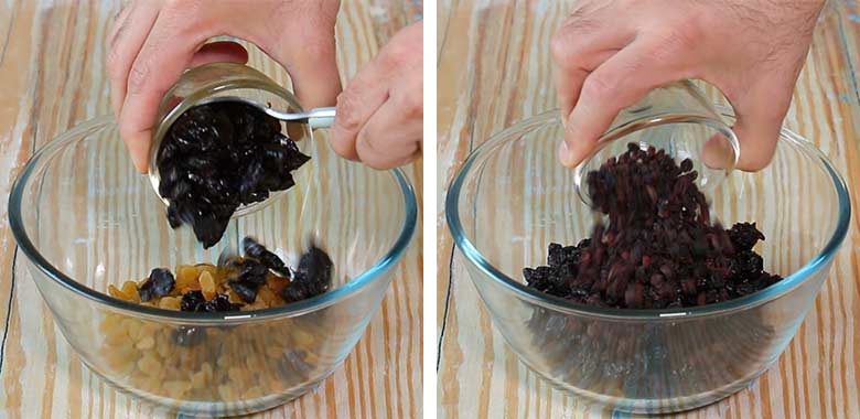 Adding prunes and black raisins to a glass bowl on a light wood table with blue staining to make Christmas Pudding