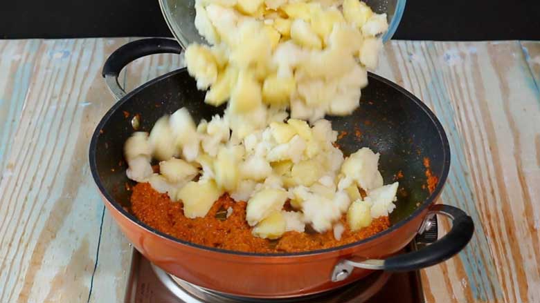 Boiled, peeled, and hand broken potatoes in a glass bowl are being tipped into an orange and black nonstick kadhai with cooked tomato masala in it to make dilli wali aloo - Delhi style Aloo Curry Recipe