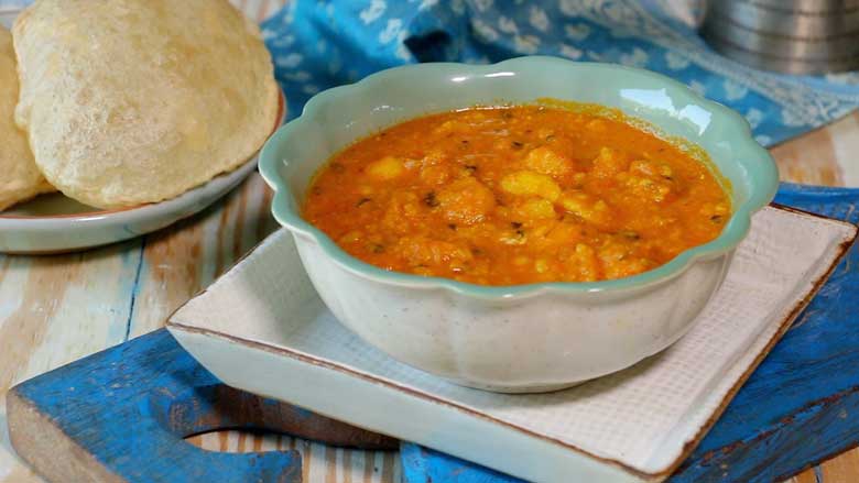 Dilli Wali Aloo are served in a jade and white fluted ceramic bowl on a white ceramic plater that has been placed on blue carved wooden piece. Freshly fried pooris and a blue tie dye textile can be seen in the background - Delhi style Aloo Curry Recipe