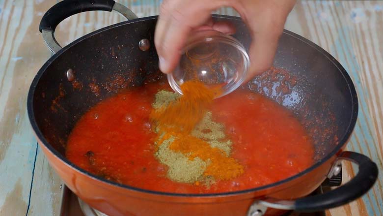 Spice powders including turmeric are being tipped into an orange and black nonstick kadhai from a small glass bowl with tomato masala being cooked in it to make dilli wali aloo -  Delhi style Aloo Curry Recipe