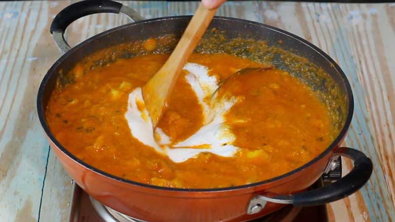 Yogurt is being stirred into the ready dilli wali aloo with a wooden spatula after the heat is turned off. The curry is in an orange and black nonstick pan with two handles on a natural wooden surface with blue streaks - Delhi style Aloo Curry Recipe