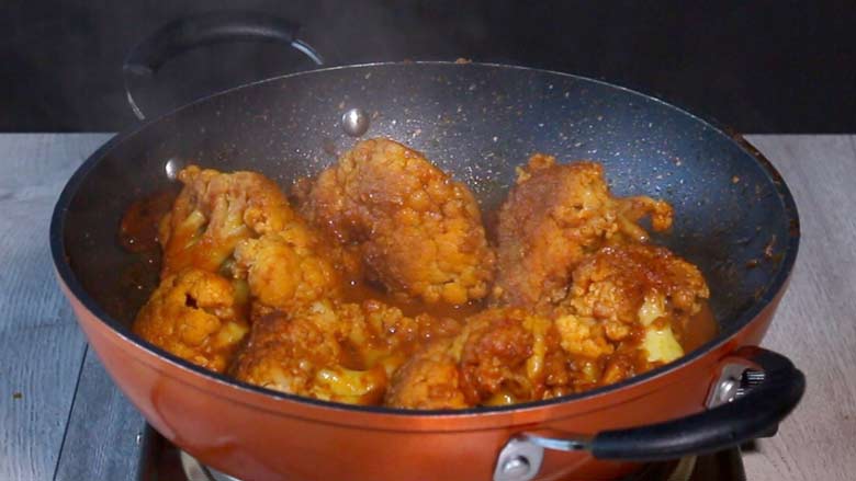 large florets of cooked gobi masala is seen in an orange and black pan with steam rising from the pan. The pan is on a stove on a grey wooden counter - Gobi Masala Recipe - Restaurant Style Cauliflower Recipe