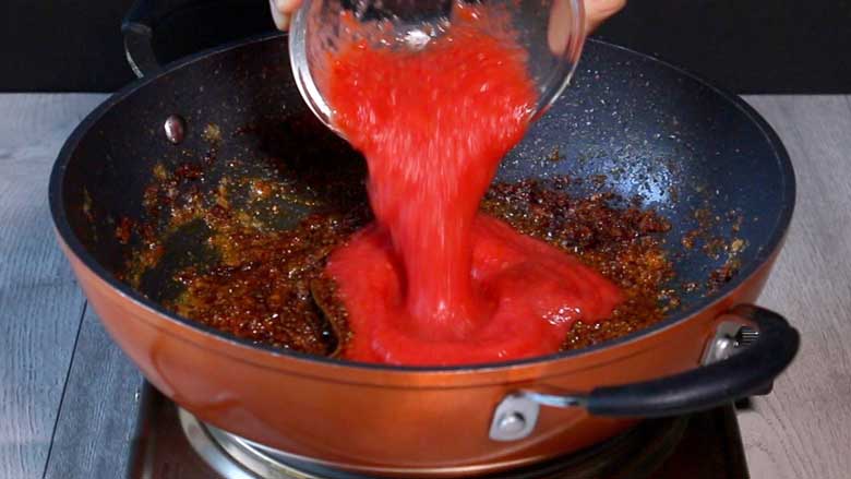 Fresh tomato puree is being poured into a orange coloured pan with sautéed onion masala that have been browned. The pan is on a metallic electric stove on a grey wooden counter top - Gobi Masala Recipe - Restaurant Style Cauliflower Recipe