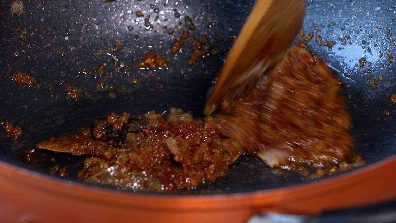 A closeup shot of onions that have been sautéed to a rich brown colour. A wooden spatula is seen stirring the onions in a black and orange non-stick pan - Gobi Masala Recipe - Restaurant Style Cauliflower Recipe