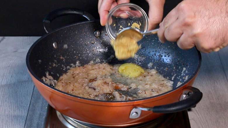 Ginger paste from a small glass bowl is being tipped into an orange and black non-stick kadhai in which onion and garlic is being sautéed - Gobi Masala Recipe - Restaurant Style Cauliflower Recipe