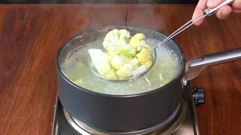 A hand is seen holding a wire ladle full of blanched cauliflower florets over a dark grey saucepan full of boiling water. The saucepan is on a metallic electric stove on a wooden surface - Gobi Shalgam Achar Recipe