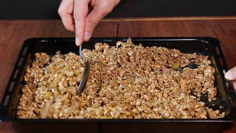 A hand is seen using a spoon to stir the baked golden brown granola in a black rectangular baking tray lying on a wooden table - Granola Recipe - Honey Banana Nut Crunch Cereal