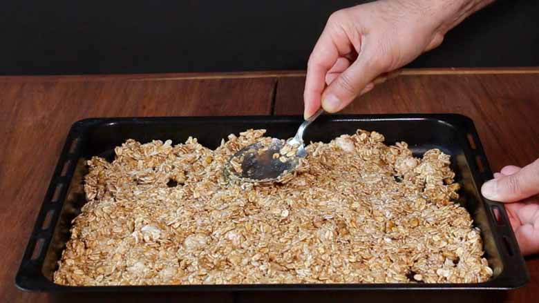 A hand is seen using the back of a spoon to spread and press down the raw granola mixture evenly in a black rectangular baking tray lying on a wooden table - Granola Recipe - Honey Banana Nut Crunch Cereal