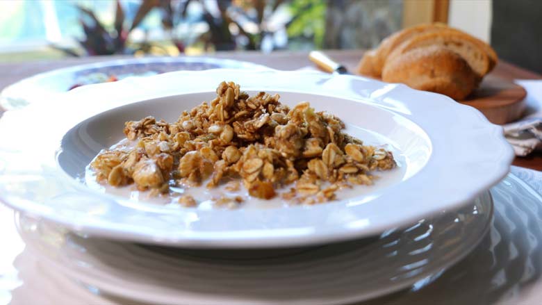 Crunchy granola with milk is seen in a close up shot in a wide white cereal bowl lying on a white plate with sliced bread and plants in the background - Granola Recipe - Honey Banana Nut Crunch Cereal