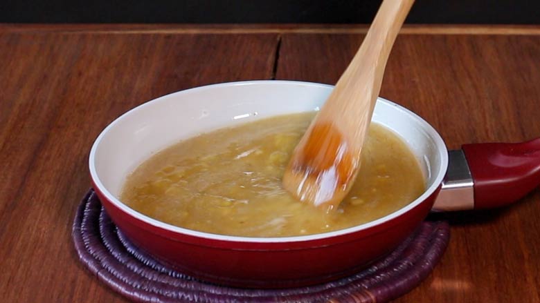 A wooden spatula is seen stirring mashed ripe banana and honey in a red frying pan with a white ceramic inner surface lying on a coloured cane mat - Granola Recipe - Honey Banana Nut Crunch Cereal