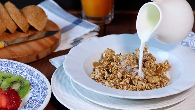 Milk is being poured from a small white ceramic milk jug over granola in a shallow white cereal bowl lying on white plates and a blue and white printed cotton mat. Sliced strawberries and kiwi fruit are seen in a blue and white plate and sliced bread and orange juice in a glass are seen in the background - Granola Recipe - Honey Banana Nut Crunch Cereal