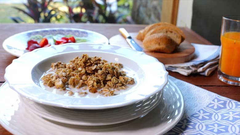 An angled side shot of granola served with milk in a shallow white cereal bowl lying on white plates and a blue and white printed cotton mat. Sliced strawberries and kiwi fruit are seen in a blue and white plate and sliced bread and orange juice in a glass are seen in the background - Granola Recipe - Honey Banana Nut Crunch Cereal