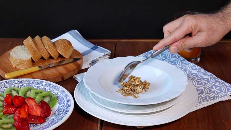 A hand is seen spooning granola into a shallow white cereal bowl lying on white plates and a blue and white printed cotton mat. Sliced strawberries and kiwi fruit are seen in a blue and white plate and sliced bread is seen in the background - Granola Recipe - Honey Banana Nut Crunch Cereal