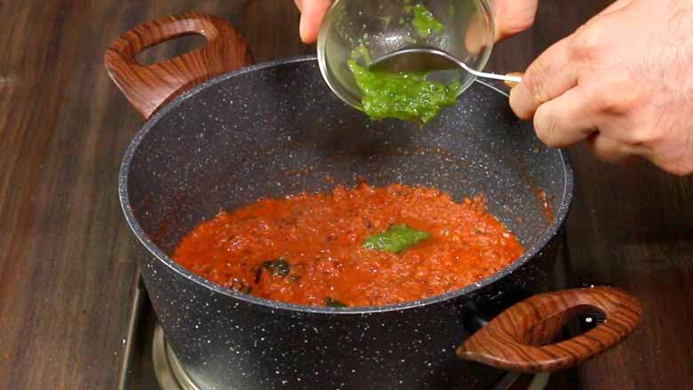 Green chilli paste is being spooned out of a small glass bowl into the cooked onion tomato mixture in a black and white speckled nonstick pan while making the egg curry - Green Chilli Egg Curry Recipe