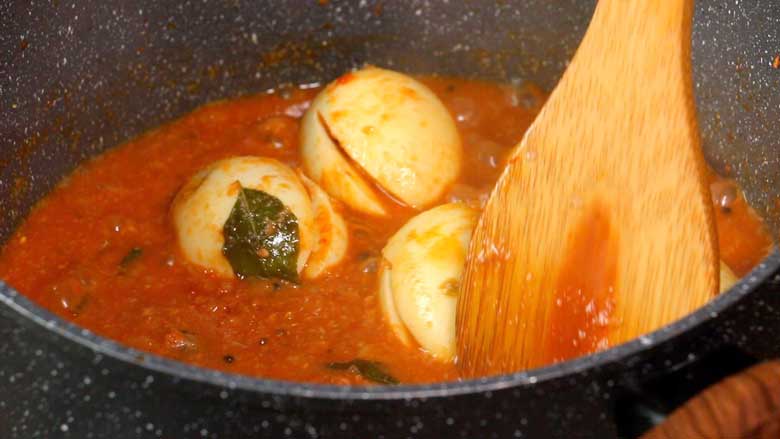 A close up shot of a wooden spatula seen gently stirring the slit full boiled eggs in the green chilli curry in a speckled black and white nonstick pan - Green Chilli Egg Curry Recipe
