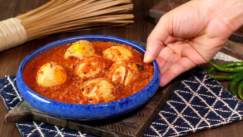 A hand is bringing a cobalt blue shallow ceramic bowl filled with green chilli egg curry and placing it on a carved wooden platter on a white and blue graphic woven fabric with green chillies and a wok brush in the background - Green Chilli Egg Curry Recipe