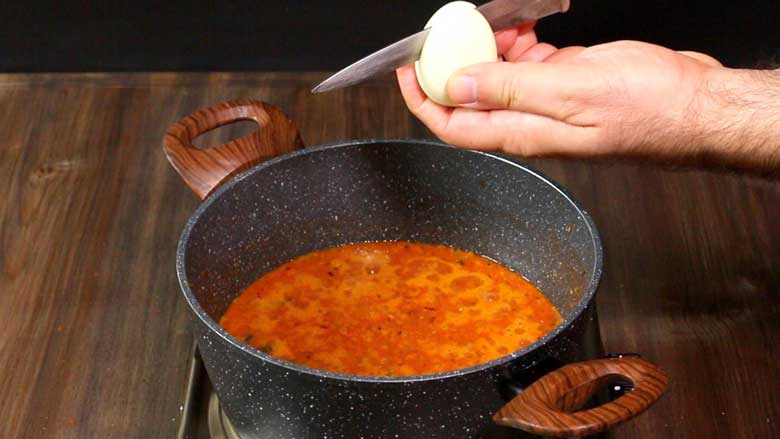 A hand is seen holding a full boiled egg that is being slit vertically from the top center over a pan of cooking onion-tomato curry in a speckled nonstick pan with two large wooden handles - Green Chilli Egg Curry Recipe