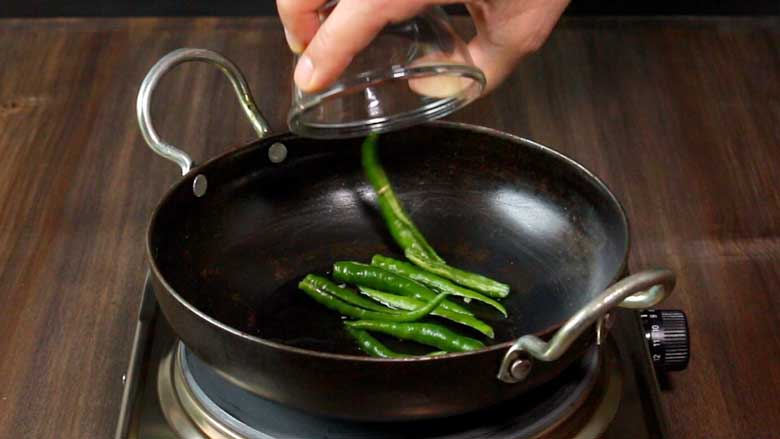 A hand is seen tipping green chillies from a small glass bowl into a small black kadhai with two handles on an electric stove on a dark wooden countertop - Green Chilli Egg Curry Recipe