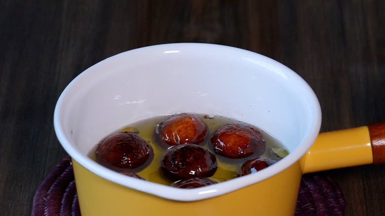 Gulab jamuns soaking in prepared sugar syrup in a yellow and white saucepan with a wooden handle on a purple straw mat on a dark wooden table - Gulab Jamun recipe with Khoya