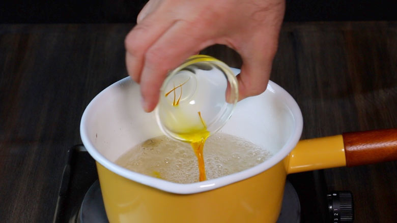A hand is seen pouring bright coloured saffron soaked in water from a small glass bowl into a white and yellow saucepan with a ceramic coating and a wooden handle with prepared sugar syrup for the gulab jamun recipe with khoya