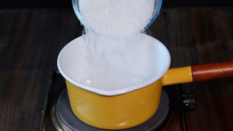 Granulated sugar being tipped from a glass bowl into hot water in a yellow and white saucepan with a ceramic coating and a wooden handle to make sugar syrup for the gulab jamun recipe with khoya