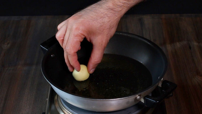 A hand is seen gently placing a gulab jamun peda or ball into hot oil in a black and silver non stick kadhai on a metallic lying on a wooden table - Gulab Jamun Recipe with khoya