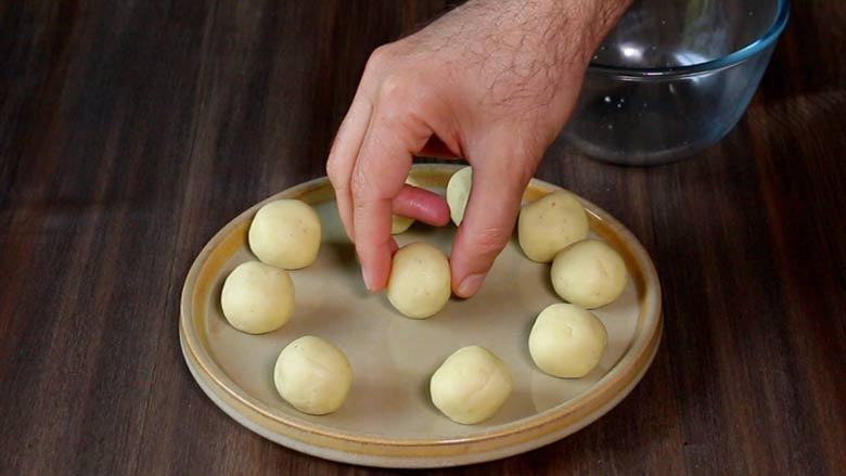 A hand is seen placing one rolled gulab jamun ball in the centre of a beige ceramic plate with nine gulab jamun dough balls already on the plate. The plate is on a dark wooden table with an empty glass bowl in the background - Gulab Jamun Recipe with khoya