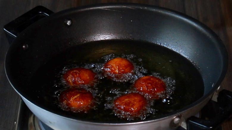A close up image of five gulab jamuns browning and frying in hot bubbling oil in a black non stick kadhai with black handles - Gulab Jamun Recipe with khoya
