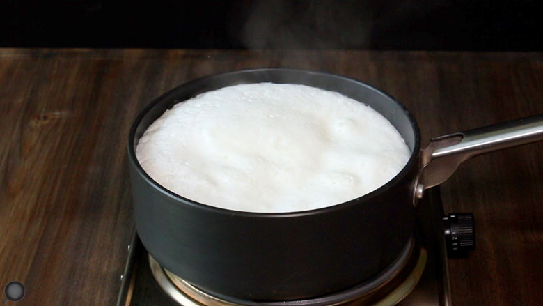 Milk boiling and rising up in a black single handled non-stick saucepan on an electric stove on a dark wood counter top - Homemade Yogurt Recipe
