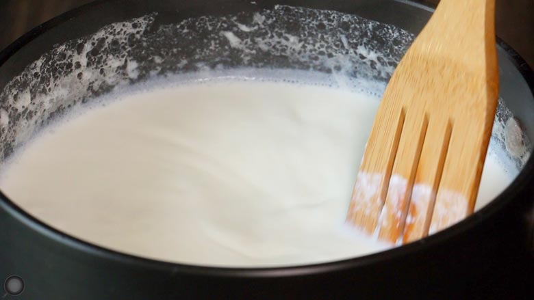 A close up image of a slotted wooden spatula stirring milk simmering in a black non stick saucepan while making dahi - Homemade Yogurt Recipe