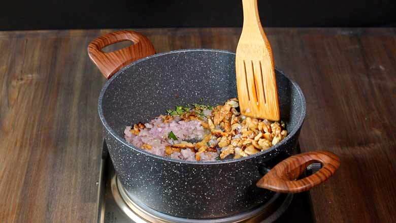 California walnuts are added and are being stirred in the onion masala by a sloth ted wooden spatula in a speckled grey and white non-stick pan with two wooden handles over a stainless steel electric stove - Indian Green beans with Walnuts Recipe Caption: Add toasted California walnuts to the pan with the onion spice mixture and sauté