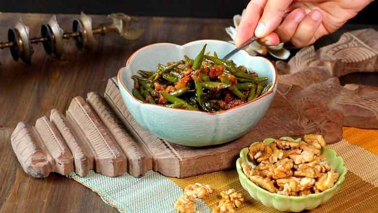 A hand is seen picking up green beans and walnuts with a spoon from a celadon blue ceramic bowl lying on a carved wooden platter. A small green bowl with California walnuts lies in the foreground Caption: Serve the Indian Green Beans with California Walnuts hot with chapati or other roti