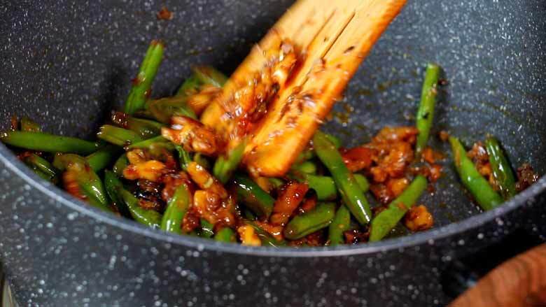A close up image of the final stages of cooking of the Indian green bean and walnuts with a wooden spatula stirring the beans in a grey and white speckled non-stick pan - Indian Green Beans and Walnuts Recipe Caption: Cook till the beans are tender