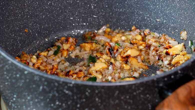 A close up shot of onions and California walnut kernels being sautéed in a speckled grey and white non-stick pan - Indian Green beans with Walnuts Recipe Caption: Sauté California walnuts and onion till the onion turns soft and translucent