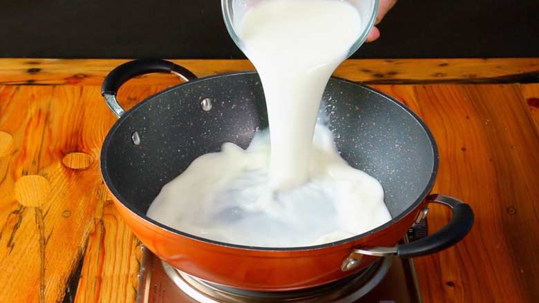 Fresh yogurt whisked with water is being poured into an orange and black nonstick kadhai or pan from a glass bowl on an electric stove on a pine wood countertop - Kashmiri Yakhni Recipe