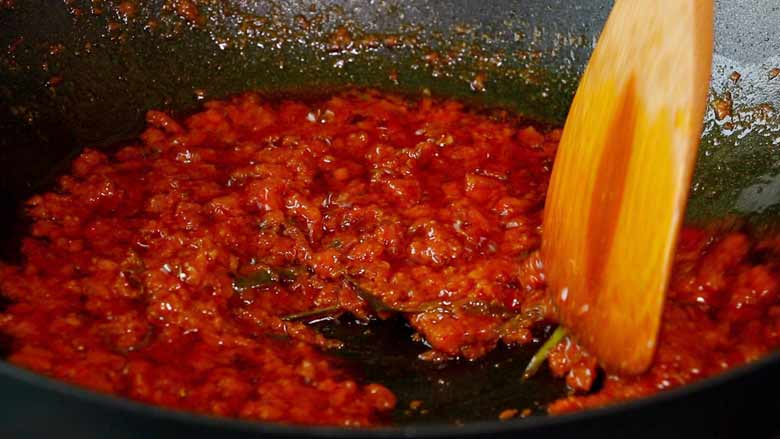 A close up image of the tomato masala being stirred by a wooden spatula in a black non stick wok. The oil can be seen separating from the masala - Kathal ki Sabzi recipe - Jackfruit Curry