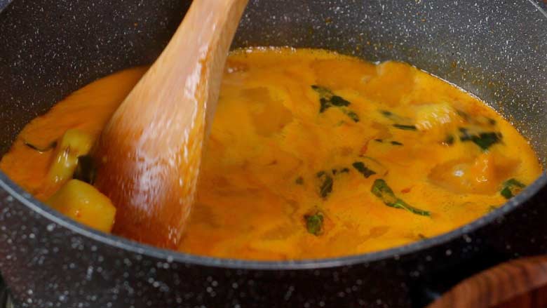 A wooden spatula is stirring Konkani potato curry that is yellow-orange in colour and has come to a boil in a black and white speckled non stick pan - Konkani Potato Curry