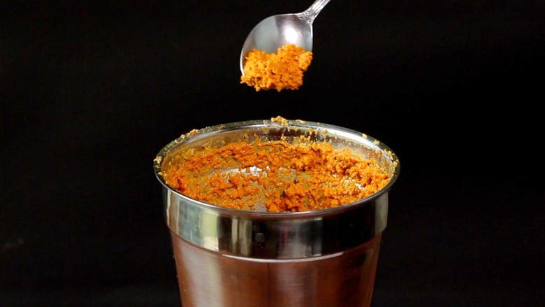 A spoon is seen picking up orange coloured ground masala paste from a steel grinder for Konkani potato curry. The image has a black background - Konkani Potato Curry Recipe