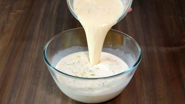 Reduced milk is being poured into the the egg custard mixture in a large glass bowl on a dark wooden countertop while making Parsi baked custard - Lagan Nu Custard Recipe
