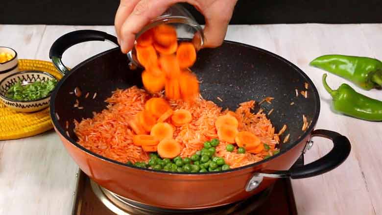 Vegetables including green peas and carrots are added to the tomato rice in an orange and black non stock pan with two handles. Two large green chillies and parsley and corn in small bowls lie in the background - Mexican Rice recipe