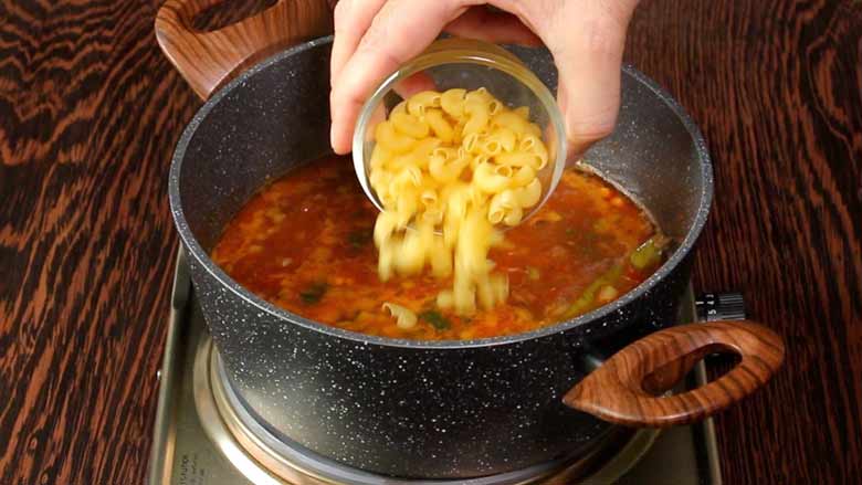 A hand is seen tipping small elbow macaroni pasta from a small glass bowl into the Italian vegetable soup cooking in a grey speckled non stick pan with two wooden handles on an electric stove on a dark wooden counter - Minestrone Soup Recipe