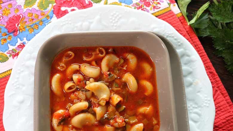 A close up top shot of minestrone soup in a grey square bowl on a white dinner plate on a red quilted tablecloth with a floral printed border with parsley stalks. The bottom of the bowl is cut off in the image - Minestrone Soup Recipe