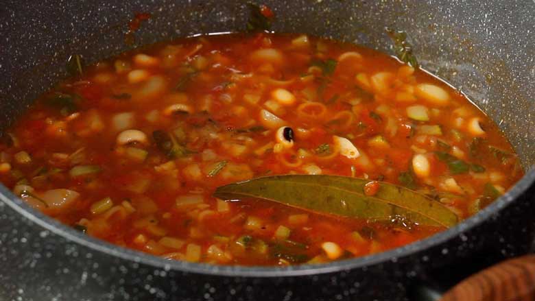 A close up shot of the cooked Italian vegetable soup in a grey speckled non stick pan with beans and bay leaf clearly seen - Minestrone Soup Recipe
