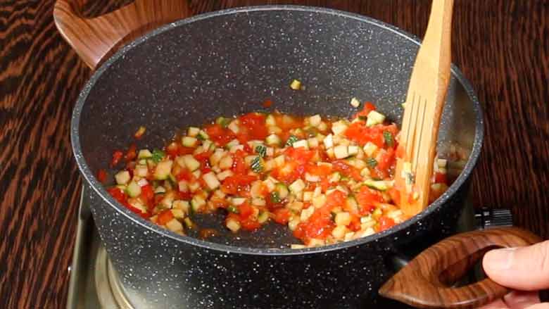 A wooden spatula is seen stirring diced vegetables in a grey speckled non stick pan with two large wooden handles on an electric stove on a dark wooden counter while making an Italian vegetable soup - Minestrone Soup Recipe