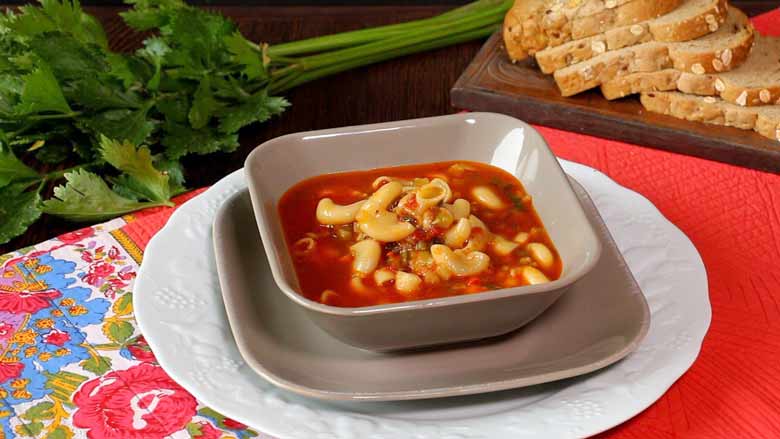 A side shot of minestrone soup served in a grey square bowl on a grey square plate lying on a white plate. The plate has been set down on a red quilted tablecloth with a floral printed border with parsley stalks and sliced bread in the background - Minestrone Soup Recipe