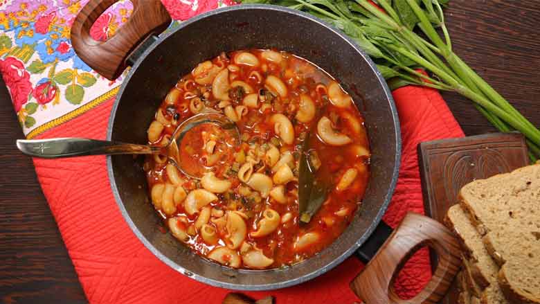 A pan with two wooden handles filled with minestrone soup with a silver ladle in it is seen lying on a red quilted tablecloth with a printed floral border. Celery stalks and bred lie in the background - Minestrone Soup Recipe