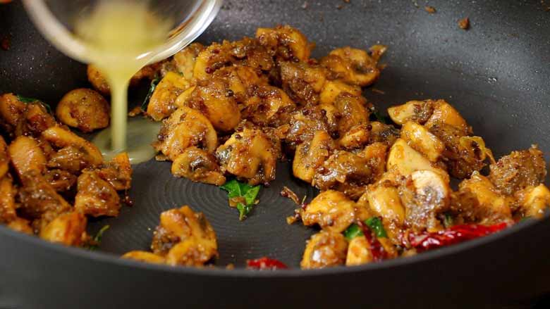 Close up image of optional ghee being added from a small glass bowl at the end of cooking mushroom pepper fry for flavour in a grey non stick pan - Mushroom Pepper Fry Recipe