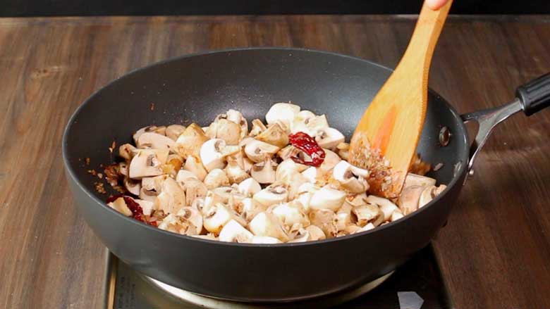 A wooden spatula stirring mushrooms that are being sautéed in a large single handled non stick pan on an electric stove on a natural wooden countertop - Mushroom Pepper Fry Recipe