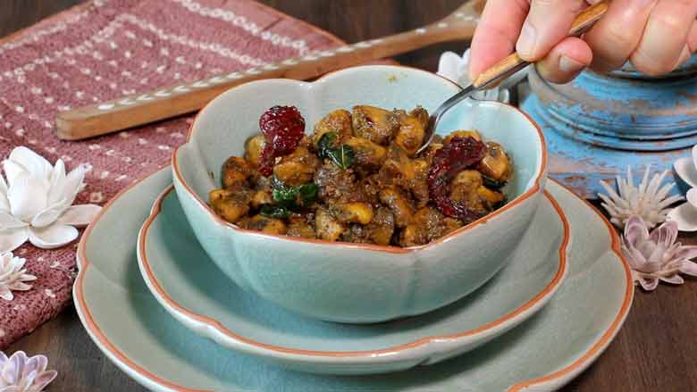 A hand is seen holding a spoon serving mushroom pepper fry from a teal ceramic bowl with a terracotta coloured rim on two plates of the same colour. A copper colour tie-dye fabric, shell flowers, inlaid wooden spoon, and a blue wooden piece lie in the background - Mushroom Pepper fry Recipe